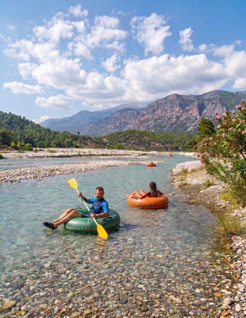 Two people sit in inflatable tubes on a shallow turquoise river, paddling with yellow oars, pebbled shore and mountains in the background.